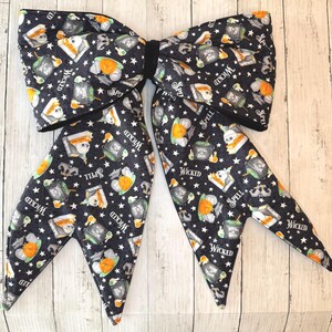 May include: A large black bow tie with a Halloween-themed pattern. The fabric features white stars, pumpkins, potion bottles, skulls, and the words "Wicked" and "Spell". The bow tie is tied with a black band.