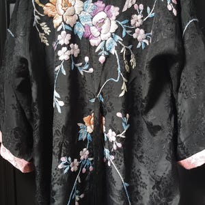 Pode incluir: Robe preta com bordados florais e de pássaros. O bordado apresenta flores em tons de pêssego, rosa e roxo, com folhas e caules azuis e verdes. A borda do robe é rosa.