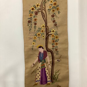 May include: A vintage embroidered wall hanging featuring a woman standing next to a tree with yellow flowers. The background is a beige fabric.