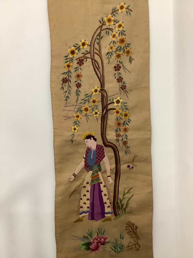 May include: A vintage embroidered wall hanging featuring a woman standing next to a tree with yellow flowers. The background is a beige fabric.