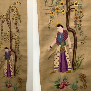 May include: A pair of embroidered wall hangings featuring a person standing next to a tree with yellow flowers. The person is wearing a colorful outfit and a crown. The background is a beige fabric.