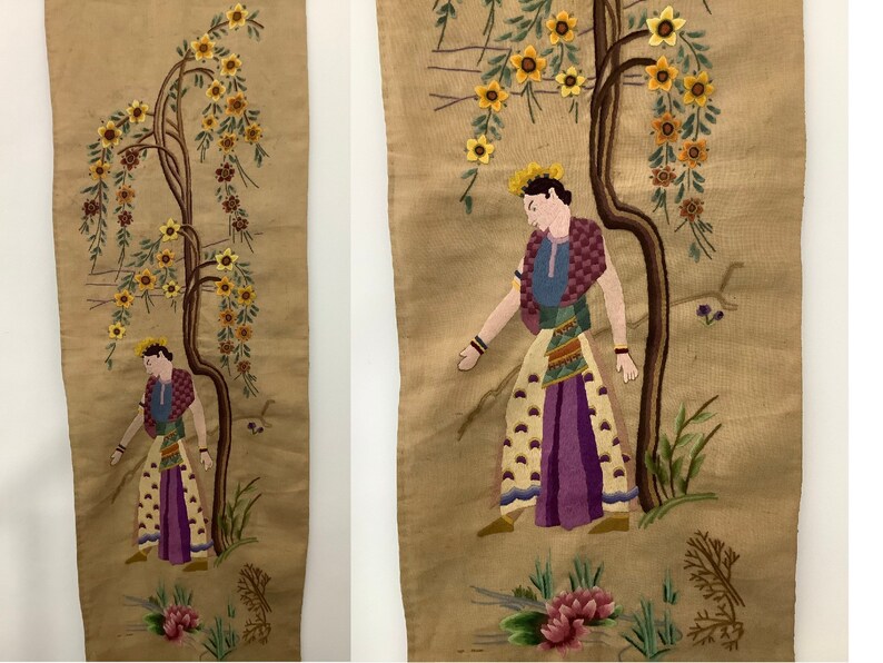 May include: A pair of embroidered wall hangings featuring a person standing next to a tree with yellow flowers. The person is wearing a colorful outfit and a crown. The background is a beige fabric.