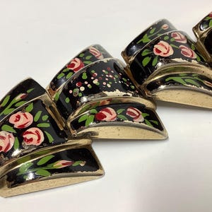 May include: A vintage bracelet featuring black enamel segments, each decorated with pink roses, green leaves, and gold-toned edges. The bracelet's design showcases a repeating floral pattern, creating a classic and elegant accessory.