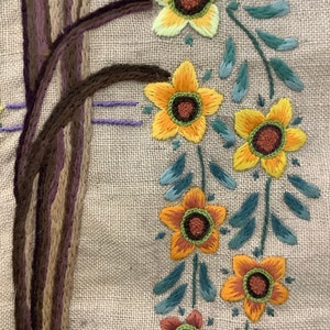 May include: Embroidered design on a beige fabric featuring a brown tree trunk and a garland of yellow, orange, and brown flowers with green leaves.