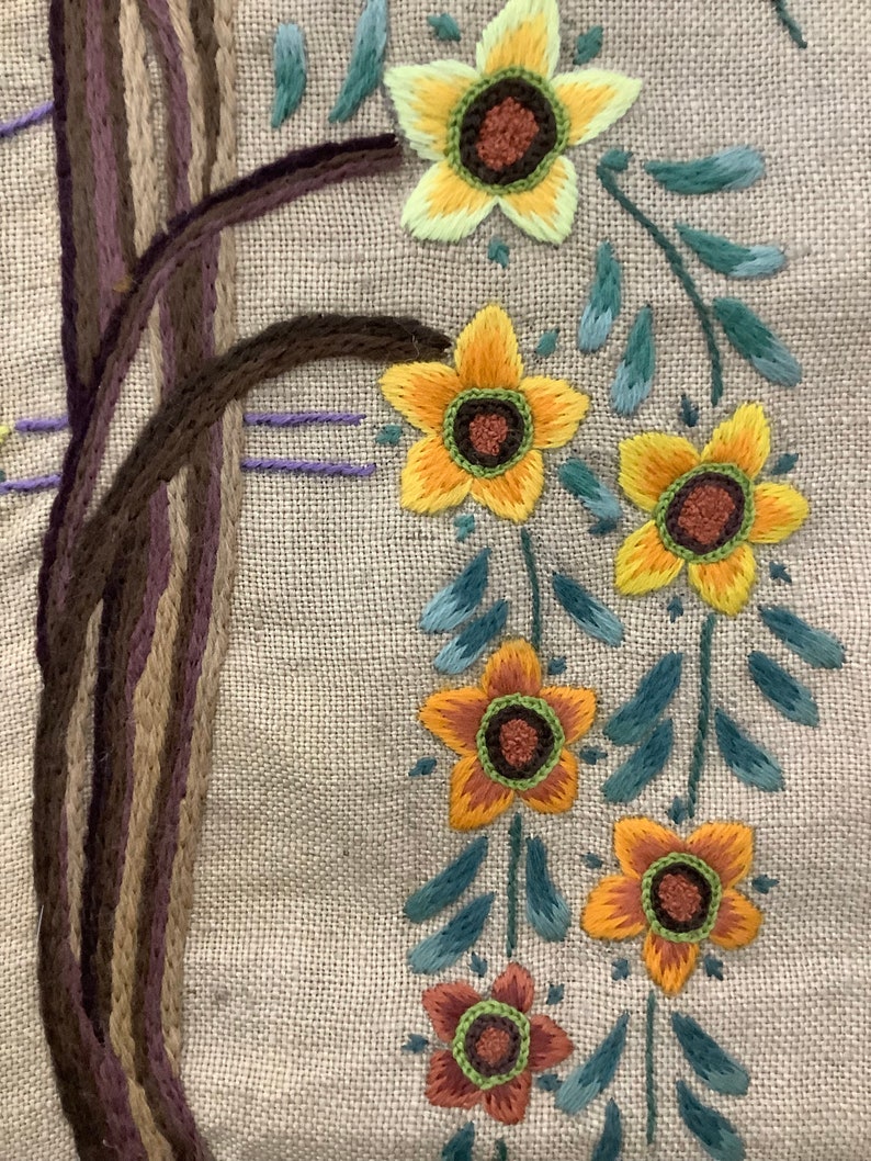 May include: Embroidered design on a beige fabric featuring a brown tree trunk and a garland of yellow, orange, and brown flowers with green leaves.