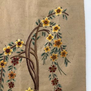 May include: A beige fabric wall hanging with embroidered flowers in shades of yellow, orange, and brown. The flowers are arranged in a floral pattern with green leaves.