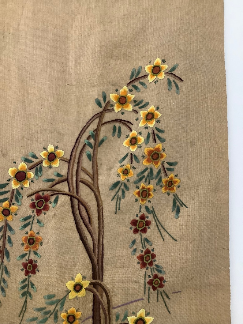 May include: A beige fabric wall hanging with embroidered flowers in shades of yellow, orange, and brown. The flowers are arranged in a floral pattern with green leaves.