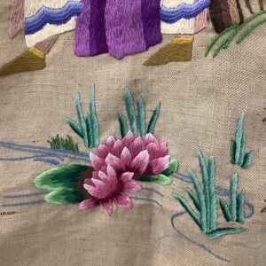 May include: Close-up of a pink embroidered flower with green leaves and stems on a beige fabric background. The flower is surrounded by blue embroidered lines that resemble a stream.