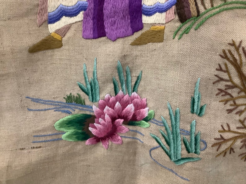 May include: Close-up of a pink embroidered flower with green leaves and stems on a beige fabric background. The flower is surrounded by blue embroidered lines that resemble a stream.