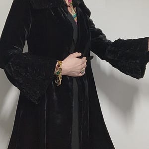 May include: A long, black velvet coat with a flared skirt and ruffled cuffs. The coat has a shawl collar and a belt at the waist. The sleeves are long and bell-shaped. The coat is a vintage style.
