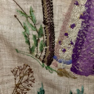 May include: Close-up of a beige linen fabric with embroidered flowers and leaves in shades of purple, pink, green, and brown. The embroidery is detailed and intricate, with a variety of stitches used.