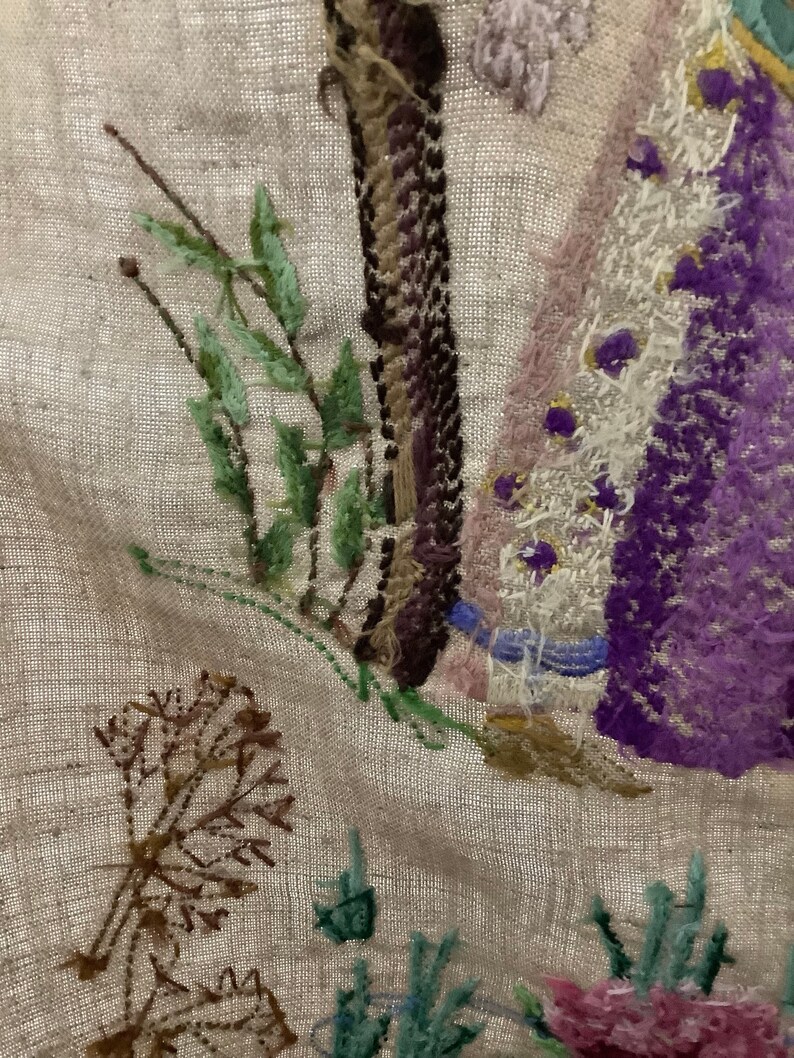 May include: Close-up of a beige linen fabric with embroidered flowers and leaves in shades of purple, pink, green, and brown. The embroidery is detailed and intricate, with a variety of stitches used.