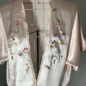 May include: A pale pink satin kimono-style jacket featuring short sleeves and a delicate lace trim along the open front edges. The garment is adorned with small, scattered floral embroidery in shades of yellow, pink, and brown across the front panels.