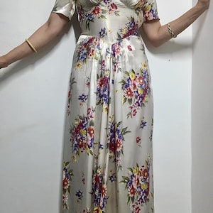 May include: A floor-length dress in a cream-coloured satin fabric featuring a vibrant floral print with red, purple, yellow, and green flowers. The dress has short sleeves, a sweetheart neckline, and a fitted bodice that transitions into a skirt.