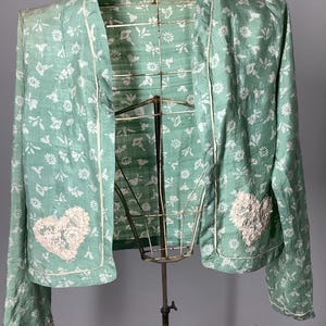 Könnte beinhalten: Eine hellgrüne Jacke mit offener Vorderseite und durchgehendem weißen Blumenmuster. Das Kleidungsstück hat lange Ärmel, weiße Paspeln an den Kanten und zwei weiße, herzförmige Spitzenapplikationen auf den unteren Vorderteilen.