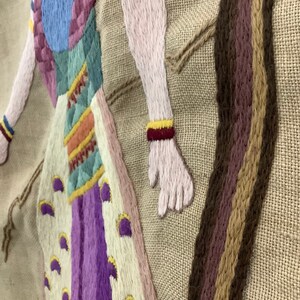 May include: Embroidered wall hanging with a colorful figure in a long dress and a long, brown braid. The figure is embroidered in shades of pink, purple, blue, yellow, and green on a beige background.