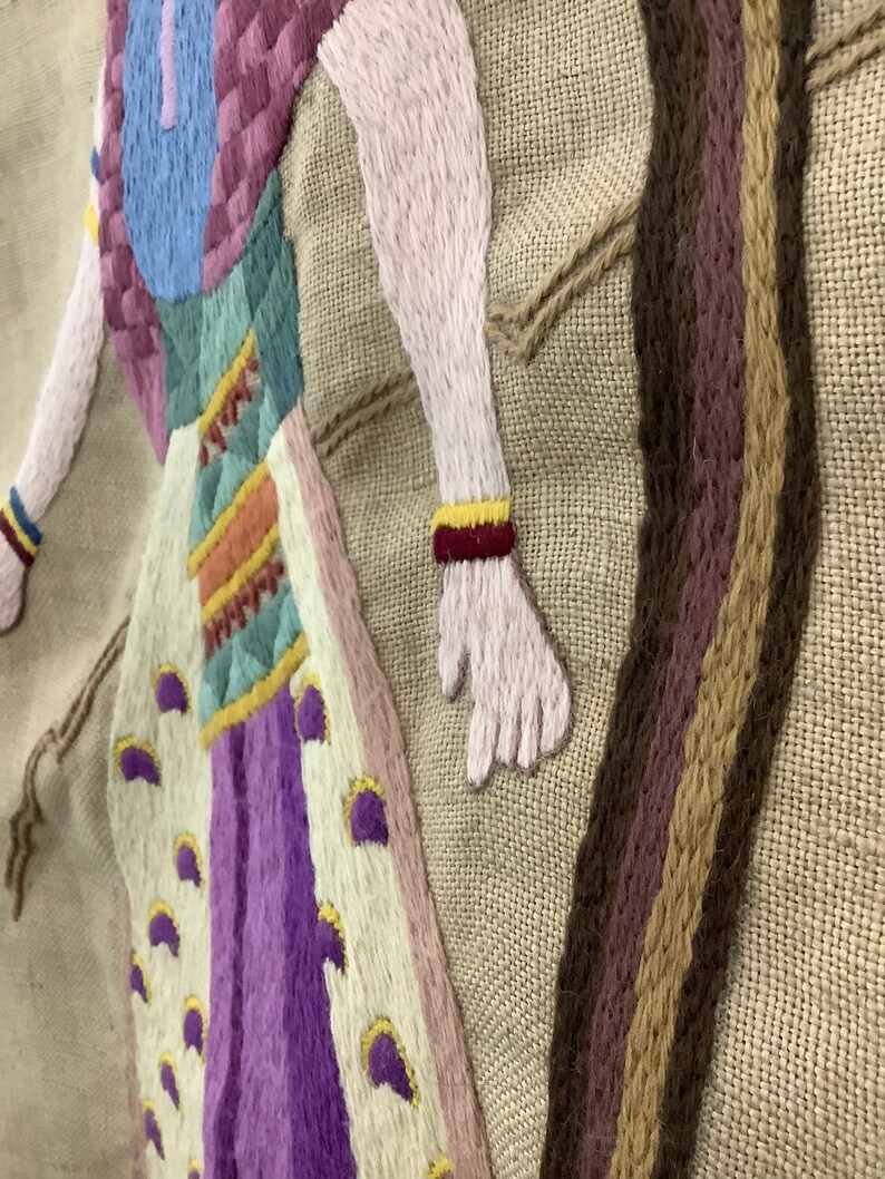May include: Embroidered wall hanging with a colorful figure in a long dress and a long, brown braid. The figure is embroidered in shades of pink, purple, blue, yellow, and green on a beige background.