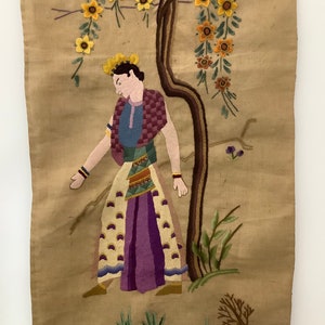 May include: Embroidered wall hanging featuring a person in a colorful outfit standing next to a tree with yellow flowers. The background is a beige fabric.