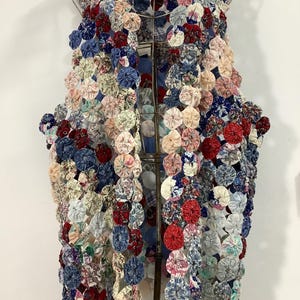 May include: A colorful vest made of fabric circles in shades of red, blue, pink, and white. The vest is displayed on a metal hanger, showcasing its open front and unique design. The fabric circles are arranged closely together, creating a textured, layered effect.