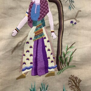 May include: Embroidered artwork depicting a person in a colorful outfit standing near a tree with flowers and leaves. The person is wearing a yellow crown, a blue shirt, a purple skirt, and a white and gold patterned sash. The background is a beige fabric.