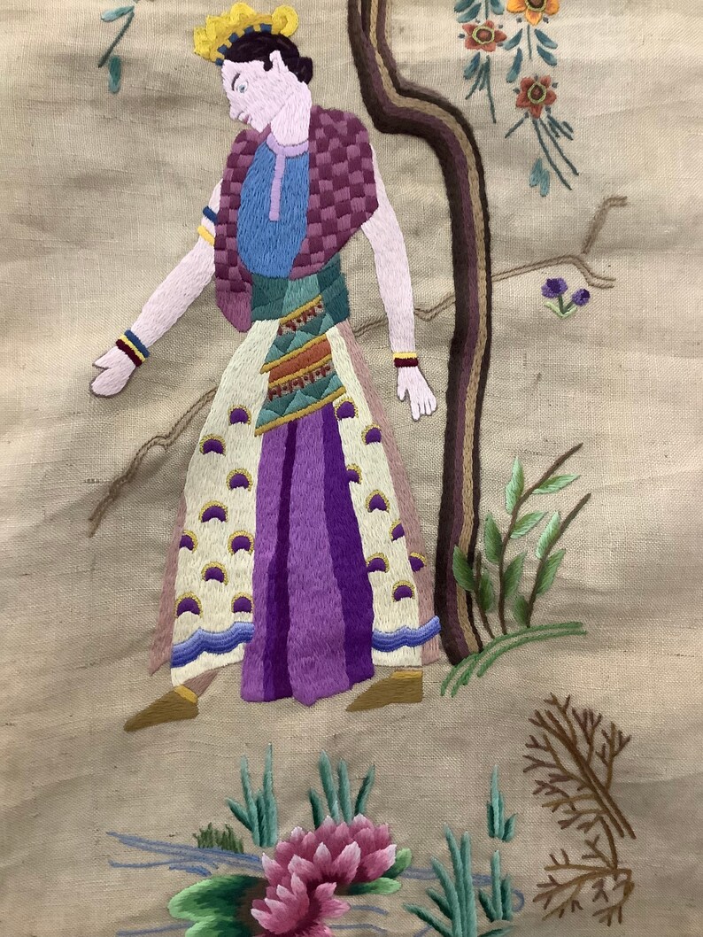 May include: Embroidered artwork depicting a person in a colorful outfit standing near a tree with flowers and leaves. The person is wearing a yellow crown, a blue shirt, a purple skirt, and a white and gold patterned sash. The background is a beige fabric.