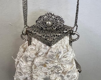 May include: A small, off-white drawstring pouch bag featuring intricate floral lace overlay. The bag is topped with a detailed, ornate silver-toned metal frame and a decorative clasp, suspended by a matching silver-toned metal chain strap.
