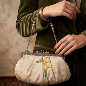 May include: A cream-colored fabric clutch bag featuring a yellow floral appliqué with green beaded stems, a silver-toned metal frame with an ornate clasp, and a delicate chain strap adorned with small beads, held against a dark olive green blazer.