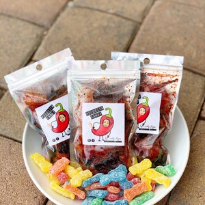 May include: Three clear plastic bags filled with red and brown spicy candy. The bags have a white label with the text "Picosita Candy" and a cartoon chili pepper with a face. A white bowl filled with colorful gummy candies.