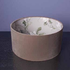 Op de afbeelding: Een beige fluwelen lampenkap met een voering met bloemenprint. De kap is cilindrisch en heeft een gladde, zachte textuur.