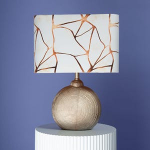 Könnte beinhalten: Eine goldfarbene Tischlampe mit strukturiertem, kugelförmigem Fuß und rechteckigem Lampenschirm. Der Lampenschirm hat einen weißen Hintergrund mit einem braunen, abstrakten geometrischen Muster. Die Lampe steht auf einem weißen, zylindrischen Ständer vor blauem Hintergrund.
