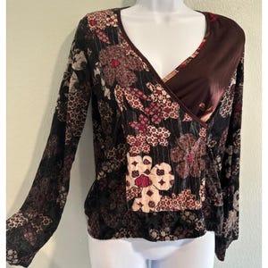 Puede incluir: Una blusa cruzada de manga larga con una mezcla de patrones florales en negro, marrón oscuro y rojo apagado. La prenda presenta un escote en V con un panel liso de color marrón oscuro en un lado y tejido texturizado con varios patrones.