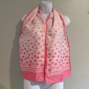 May include: A pink and white scarf with a polka dot and striped pattern. The scarf has a solid pink border and is draped over a white mannequin. The scarf is made of a lightweight material.