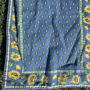May include: A blue and yellow floral patterned textile. The textile features a repeating pattern of green and white leaf-like shapes on a blue background. The border is decorated with yellow sunflowers and green leaves.