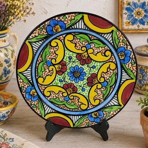 Peut inclure: Une assiette décorative en céramique avec un motif floral vibrant. L'assiette présente un bord noir, une bordure bleue et un motif central de fleurs jaunes, vertes et rouges. Elle est présentée sur un support noir.