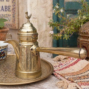 Peut inclure: Une cafetière en laiton vintage avec une poignée allongée et un ornement en forme d'oiseau sur le couvercle, posée sur un plateau en laiton. La cafetière est gravée de motifs. Deux petites tasses à motifs sont à proximité. Le fond indique "CAFE ARABE".