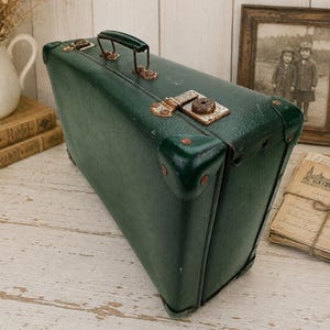 Peut inclure: Une valise vintage vert foncé avec un extérieur texturé, dotée d'une poignée supérieure, de coins renforcés et de fermoirs métalliques présentant des signes visibles d'oxydation. La valise rectangulaire a une structure rigide.