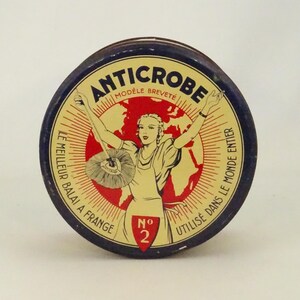 Peut inclure: Une boîte ronde en métal bleu et rouge avec les mots "ANTICROBE" et "MODÈLE BREVETÉ" imprimés dessus. La boîte représente une femme en robe jaune tenant un balai avec le texte "LE MEILLEUR BALAI A FRANGE" et "UTILISÉ DANS LE MONDE ENTIER" imprimé autour de la femme. Le numéro "2" est imprimé dans un cercle rouge sur la boîte.