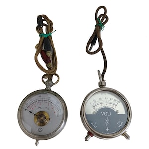 Puede incluir: Dos voltímetros antiguos con esferas circulares y carcasas metálicas. Uno tiene una esfera blanca con "VOLTS" y un indicador de aguja. El otro tiene una esfera gris con "VOLT" y una aguja. Ambos tienen cables y terminales adjuntos.