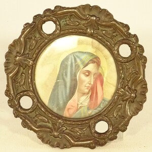 May include: A round, ornate, gold-toned metal frame with a circular image of the Virgin Mary inside. The frame has a decorative design with floral motifs and four holes around the edge.