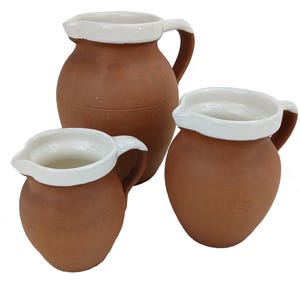 May include: Three terracotta jugs of different sizes. Each jug has a white rim and handle. The jugs are a warm brown colour, with white accents providing contrast. The jugs are likely for decorative or functional use.