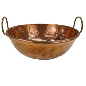 May include: A copper bowl with two brass handles. The bowl has a wide, shallow shape and a hammered texture. The copper has a warm, reddish-brown color. The handles are curved and attached to the sides of the bowl.