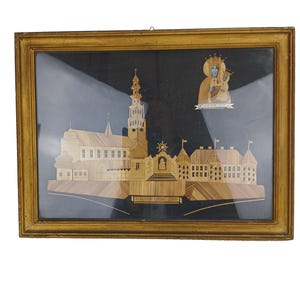 Puede incluir: Obra de arte enmarcada que presenta una detallada representación de una iglesia y edificios, elaborada con tiras de madera de color claro sobre un fondo negro. La obra está encerrada en un marco dorado, con un icono religioso en la esquina superior derecha.