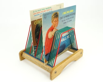 Può includere: Un porta dischi in legno con divisori in metallo rosso, blu e verde contiene una collezione di dischi in vinile vintage. Le copertine dei dischi presentano testo in francese e immagini di musicisti, tra cui "L'Idole des Jeunes" e "John Hallyday".