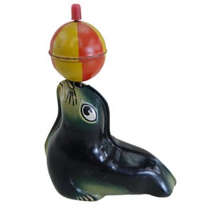 Könnte beinhalten: Vintage Spielzeug Robbe, die einen Ball balanciert. Die Robbe ist dunkelgrün und schwarz mit einem gelb-roten Ball. Das Spielzeug hat einen roten Knopf auf dem Ball.