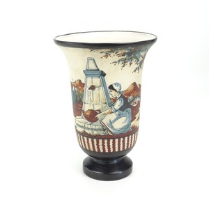 May include: A white ceramic vase with a black rim and base. The vase is decorated with a hand-painted scene of a woman drawing water from a well. The scene is painted in shades of brown, blue, and red.
