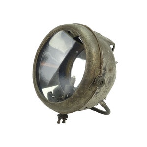 May include: A vintage metal motorcycle headlight with a clear glass lens and a rusty patina. The headlight has a round shape and a metal bracket for mounting.