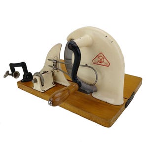 Peut inclure: Une trancheuse à viande ancienne de couleur crème avec une poignée et une lame noires, montée sur une base en bois. La trancheuse comporte une poignée en bois et une pince. Un logo rouge et blanc est visible sur le côté.