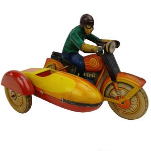 May include: Vintage toy motorcycle with sidecar. The motorcycle is painted in shades of orange, yellow, and red, with a driver figure in green and blue. The sidecar is yellow and red. The toy has a classic, aged appearance.