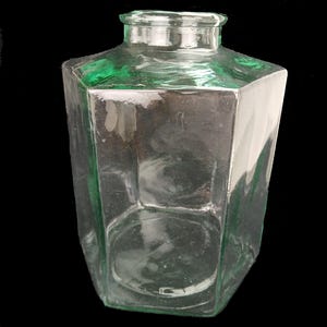 Può includere: Una bottiglia di vetro trasparente e sfaccettata con una sfumatura verde. La bottiglia ha una forma rettangolare con lati angolati e un collo stretto. Il vetro ha una superficie leggermente strutturata, su sfondo nero.