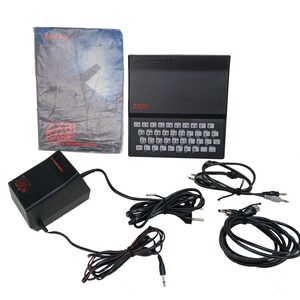 Könnte beinhalten: Ein schwarzer Sinclair ZX81-Computer mit Tastatur. Das Bild zeigt ein Netzteil, Kabel und ein Handbuch mit dem Titel "ZX81 BASIC". Der Computer hat ein schlichtes Design, das sich auf die Funktionalität konzentriert.